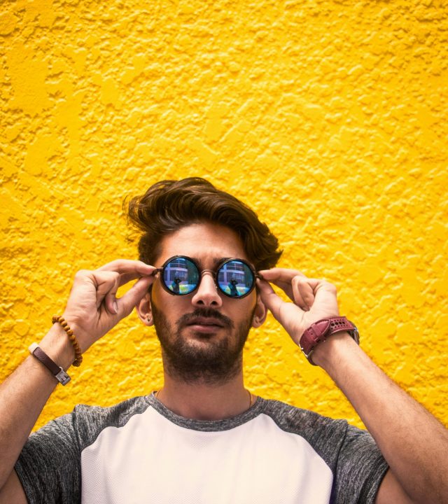 Young man with sunglasses standing against a vibrant yellow wall in daylight.
